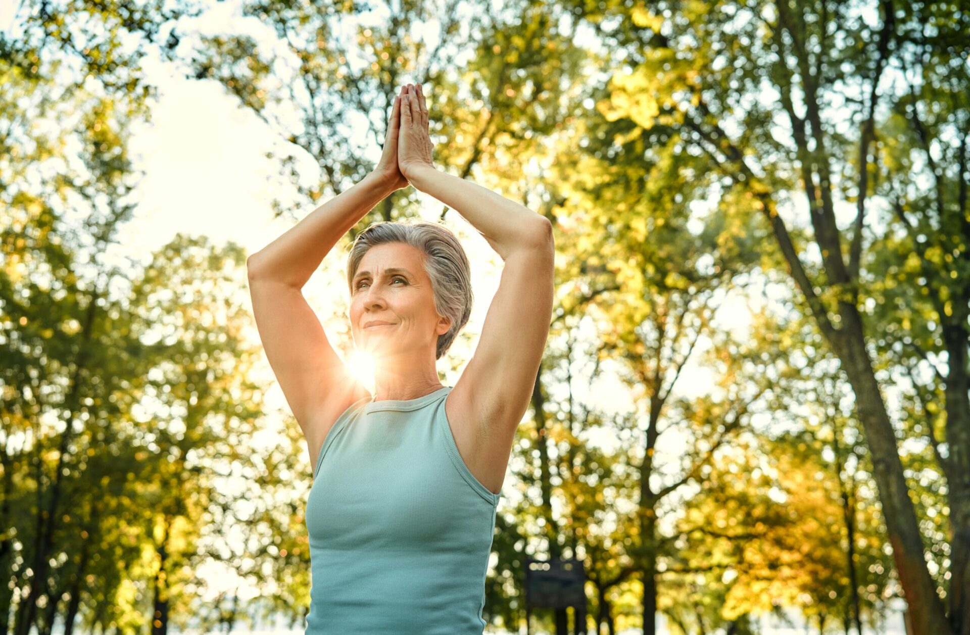 A women over 60 holds her hands together over her head against a sunny forest background